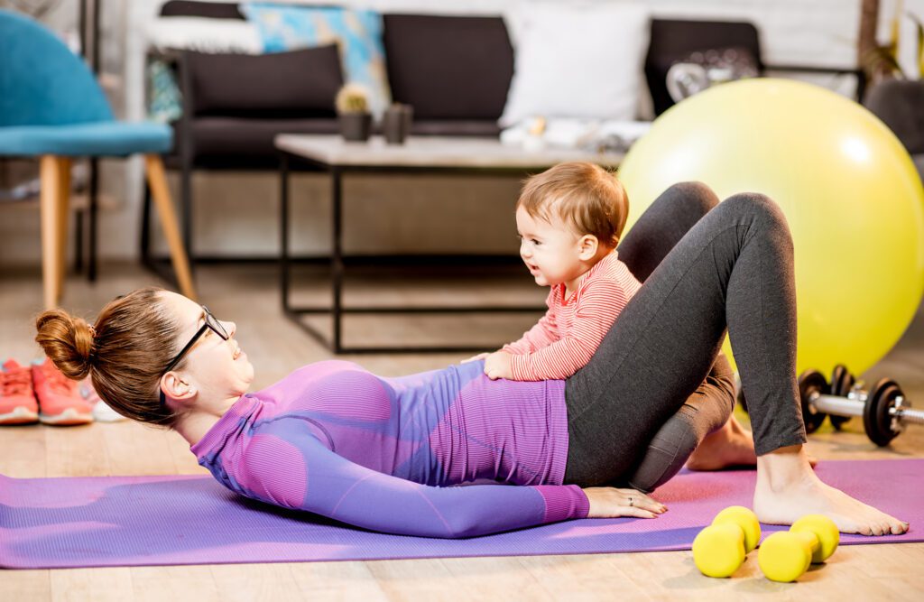 Young mother in sportswear doing exercise with her playful baby son lying on the mat at home