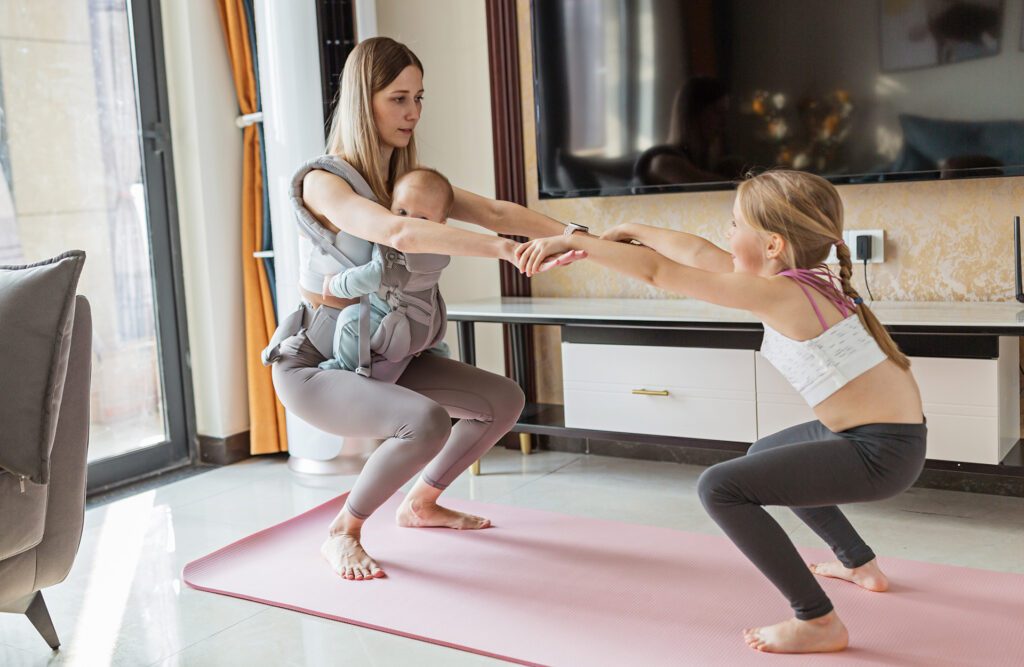 Woman stretching at home with her daughter and a baby strapped to her chest