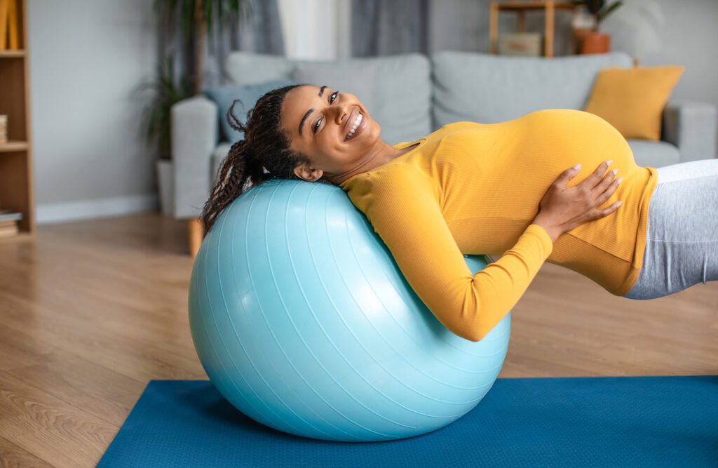 Happy young pregnant woman touching big belly, doing exercises for back on fit ball